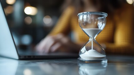 A clear glass hourglass with sand flowing down is placed next to a person working on a laptop, symbolizing the essence of time management and productivity in a modern workspace.